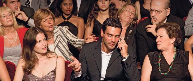 A man on a cell phone in a theater, surrounded by annoyed audience members.