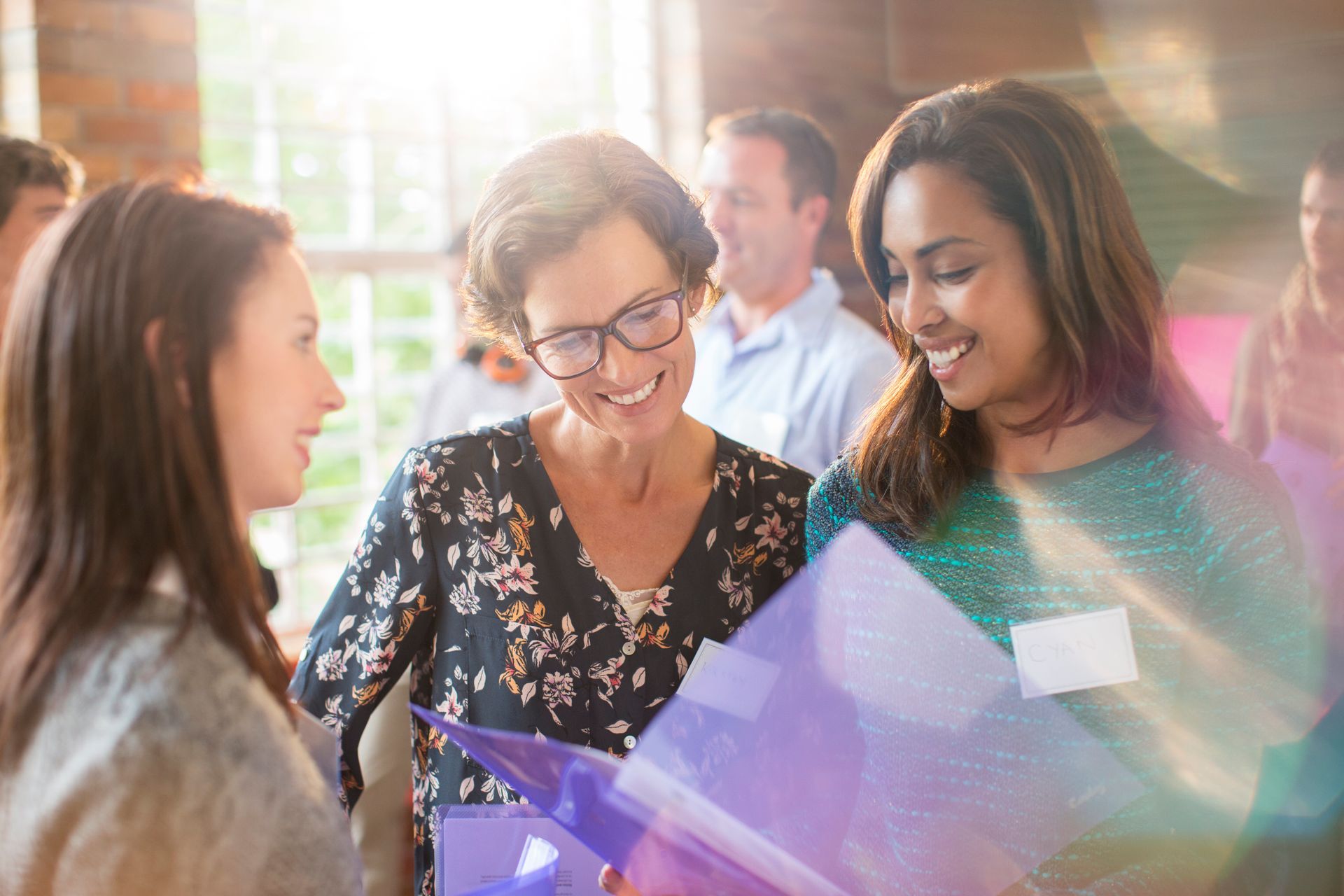 Three women looking at a purple binder, smiling, in a room with other people.