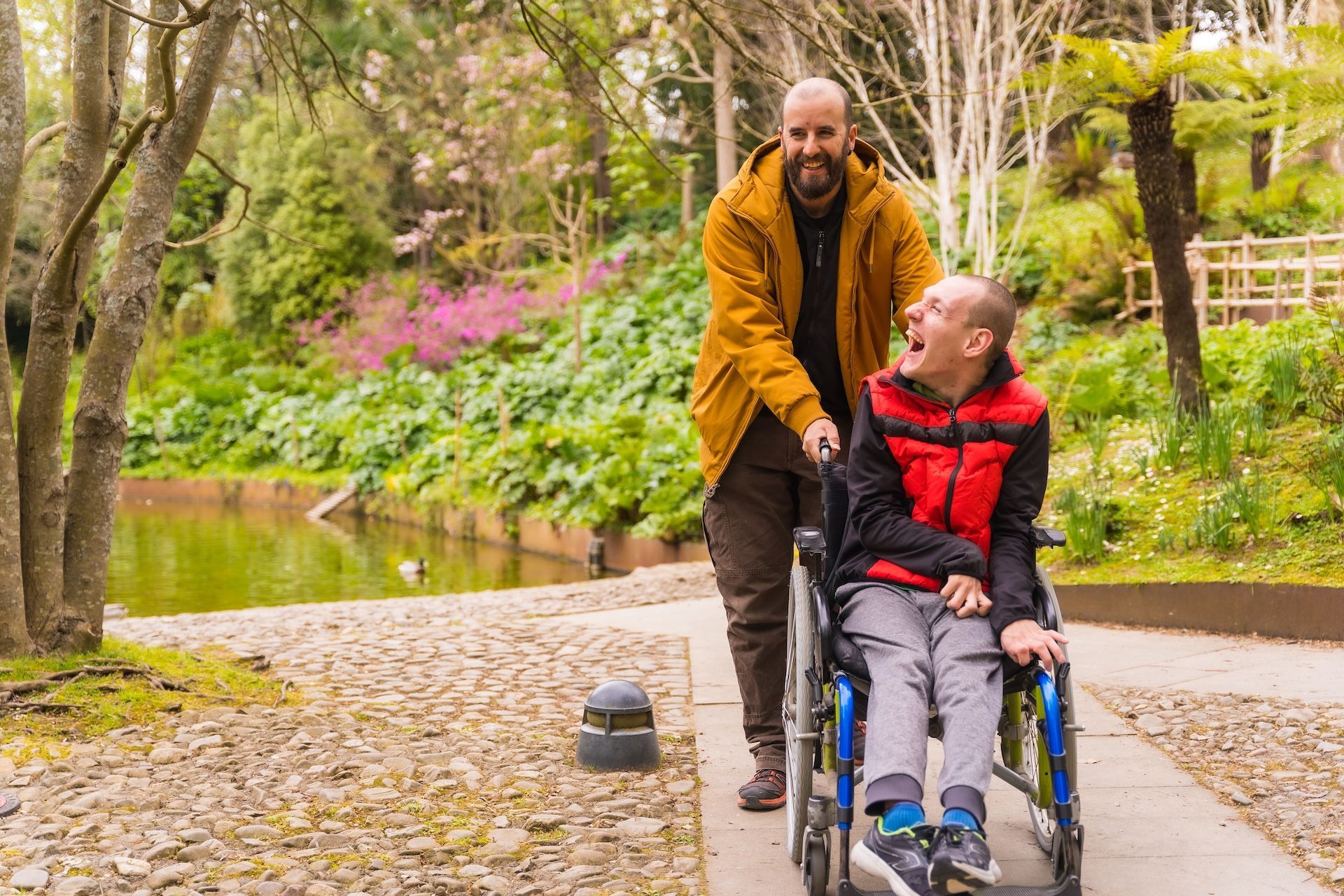 Man pushes person in wheelchair along a path in a park; both smiling.