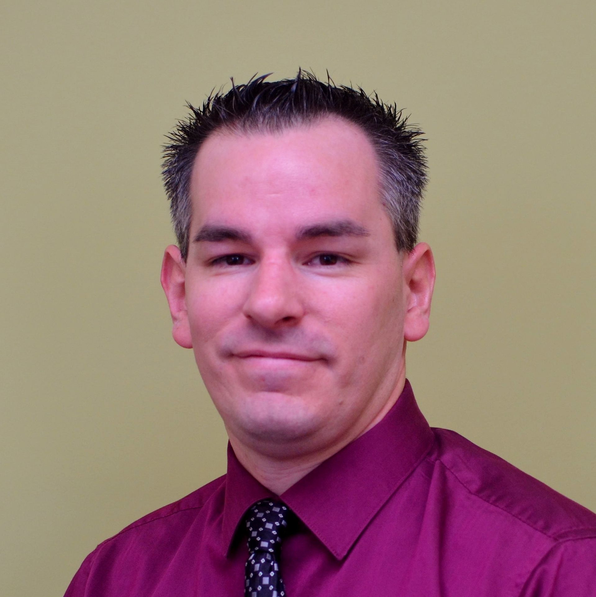 Man in burgundy shirt and patterned tie smiles, against a light green wall.