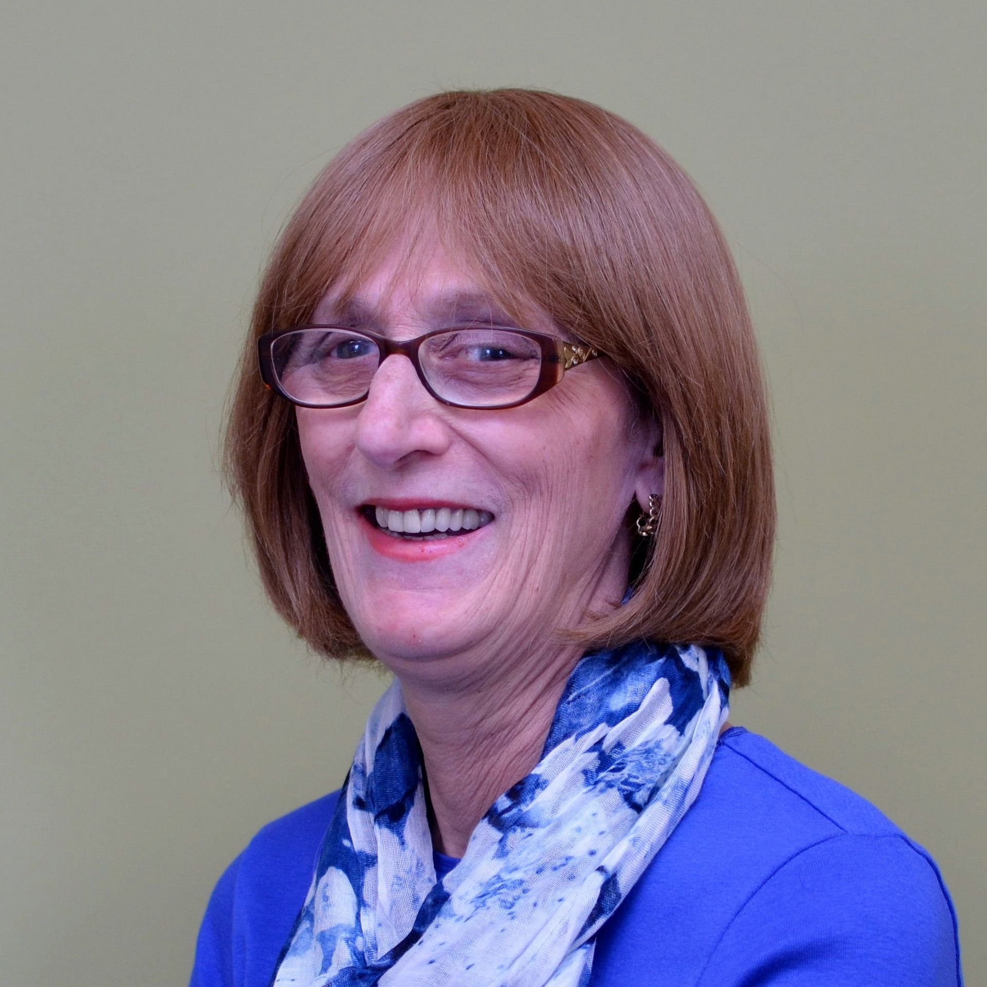 Woman with glasses smiles; wearing a blue blazer and scarf with a light brown wig.