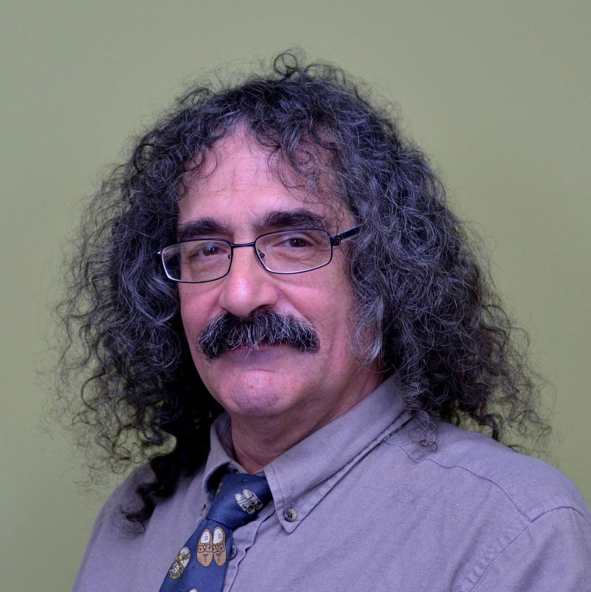 Man with curly gray hair, glasses, mustache, wearing a button-down shirt and tie, looking at the viewer.