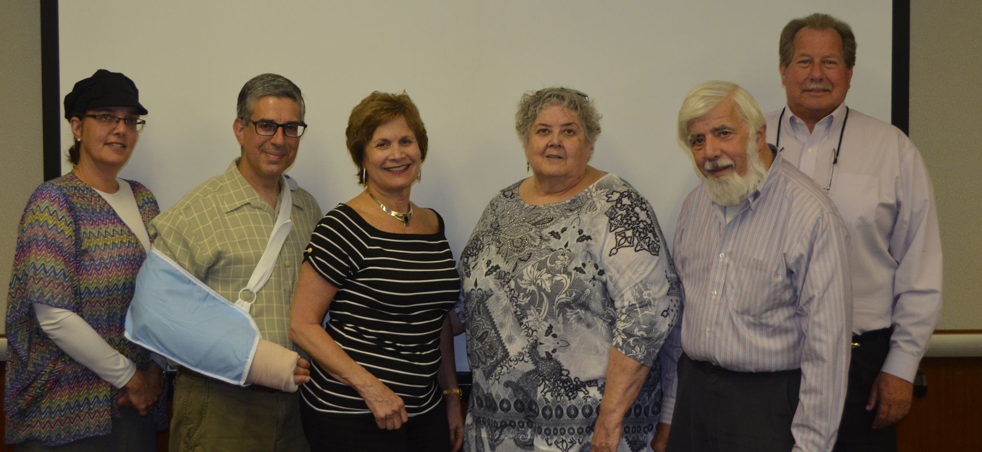 Group of seven people smiling in front of a white screen, some wearing glasses, one with an arm sling.