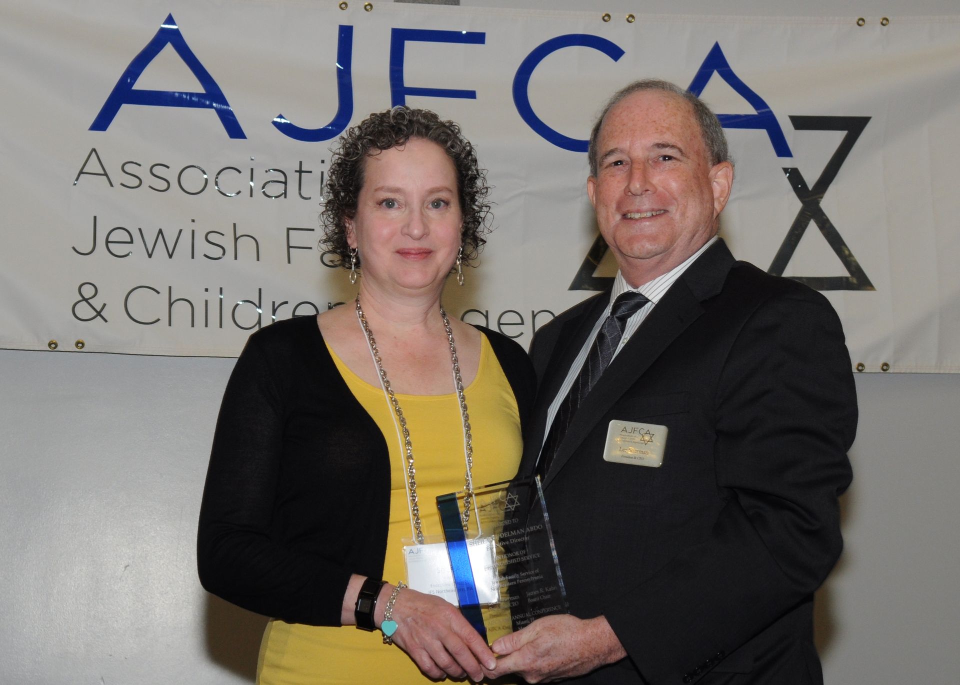 A woman in yellow and black jacket accepts an award from a man in a suit in front of an AJFCA banner.