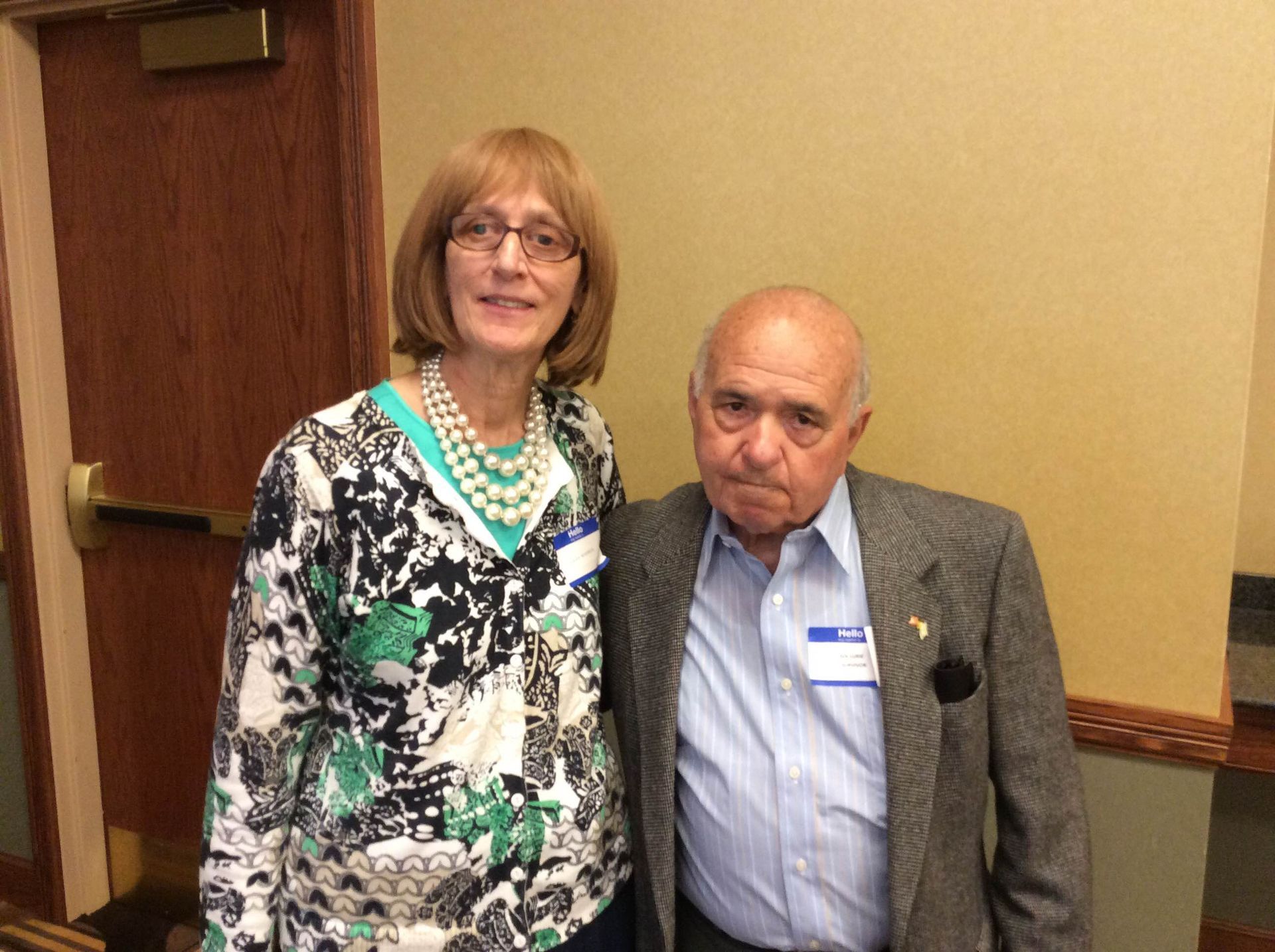 A woman with glasses and a floral shirt poses with an older man in a blazer; both have name tags.
