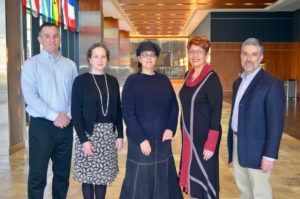 Five people standing indoors, posed. Flags in background.