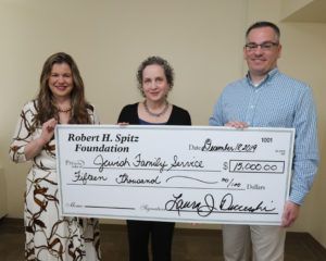 Three people hold a check for Jewish Family Service, donation for $15,000.  They are smiling in a room.