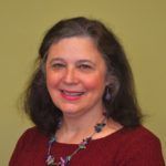Woman with brown hair smiles, wearing a burgundy sweater and a beaded necklace. Background is a solid green-yellow.
