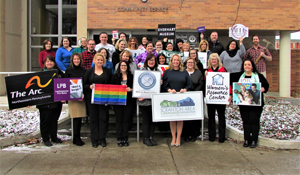 Group of people holding signs in front of a building with “Library Museum” sign. Some signs have logos/text. Snowy ground.