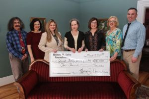 Group of people holding a large check, posing on a red sofa in a room with paintings on the wall.