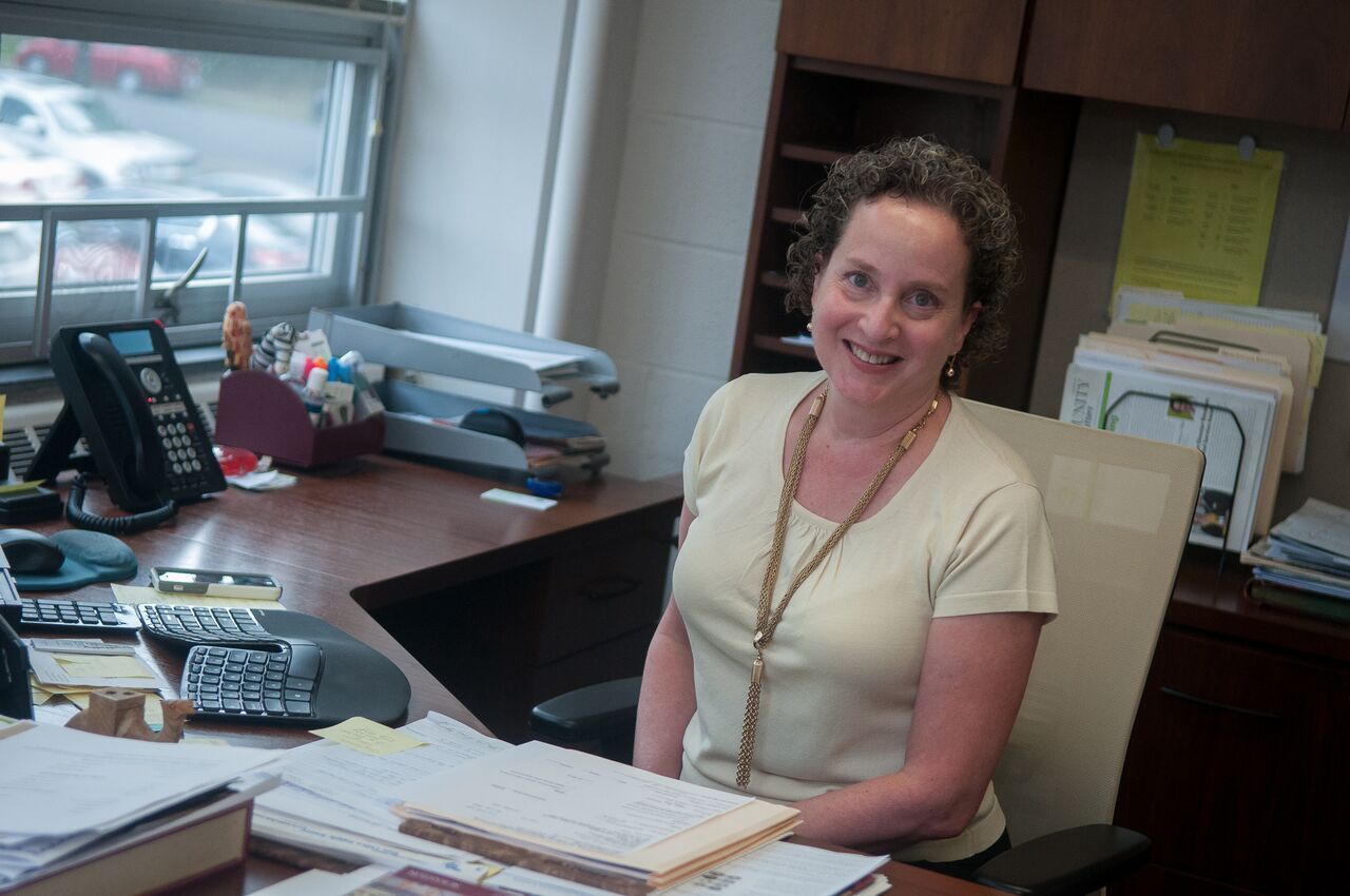 Woman smiling at her desk in an office. Beige top, brown necklace, papers, phone, and window visible.