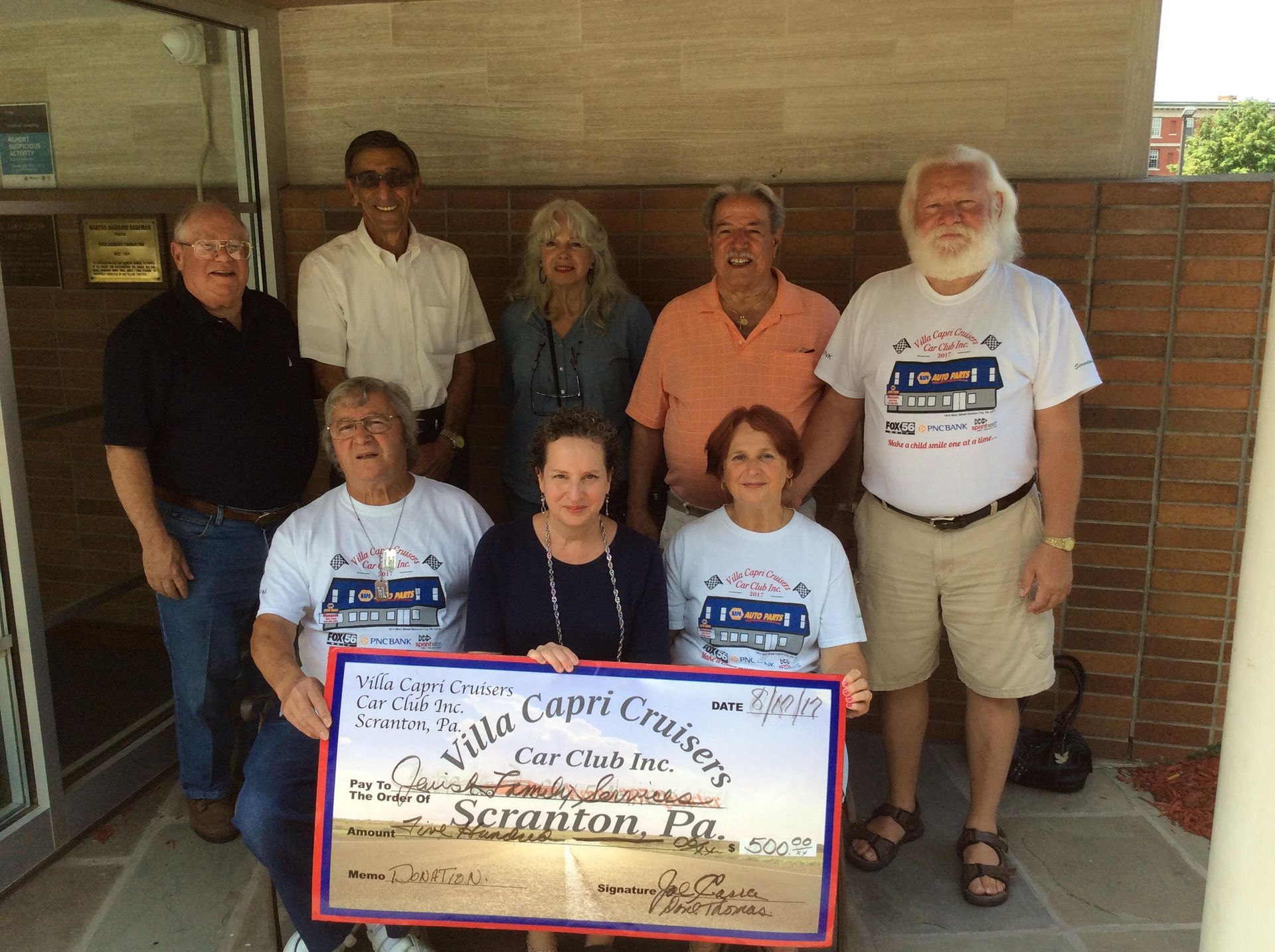 Group of people holding a check in front of a building. Capri Cruisers donation to Scranton, PA.