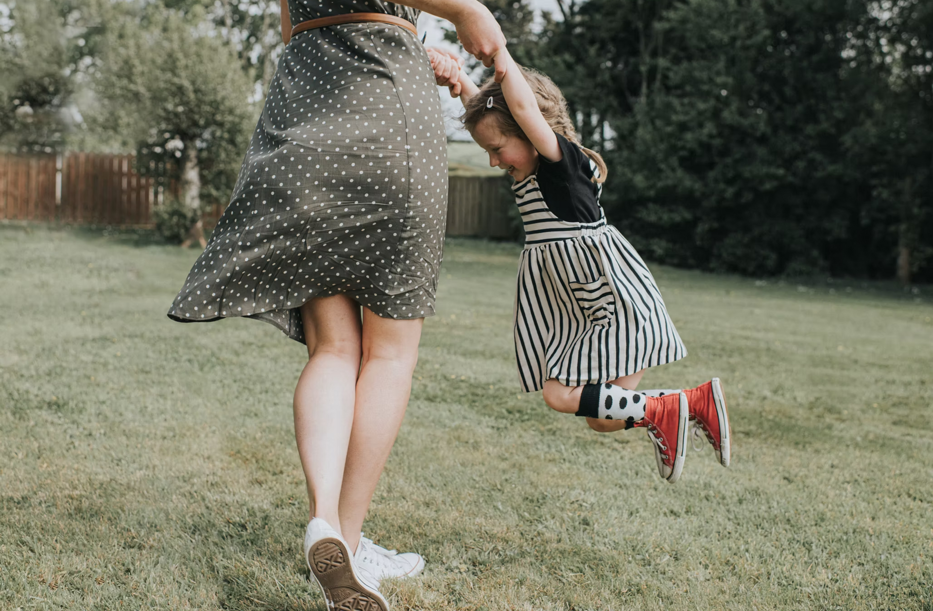 Woman swings child by the hands across a grassy backyard; child wears striped dress and red shoes.