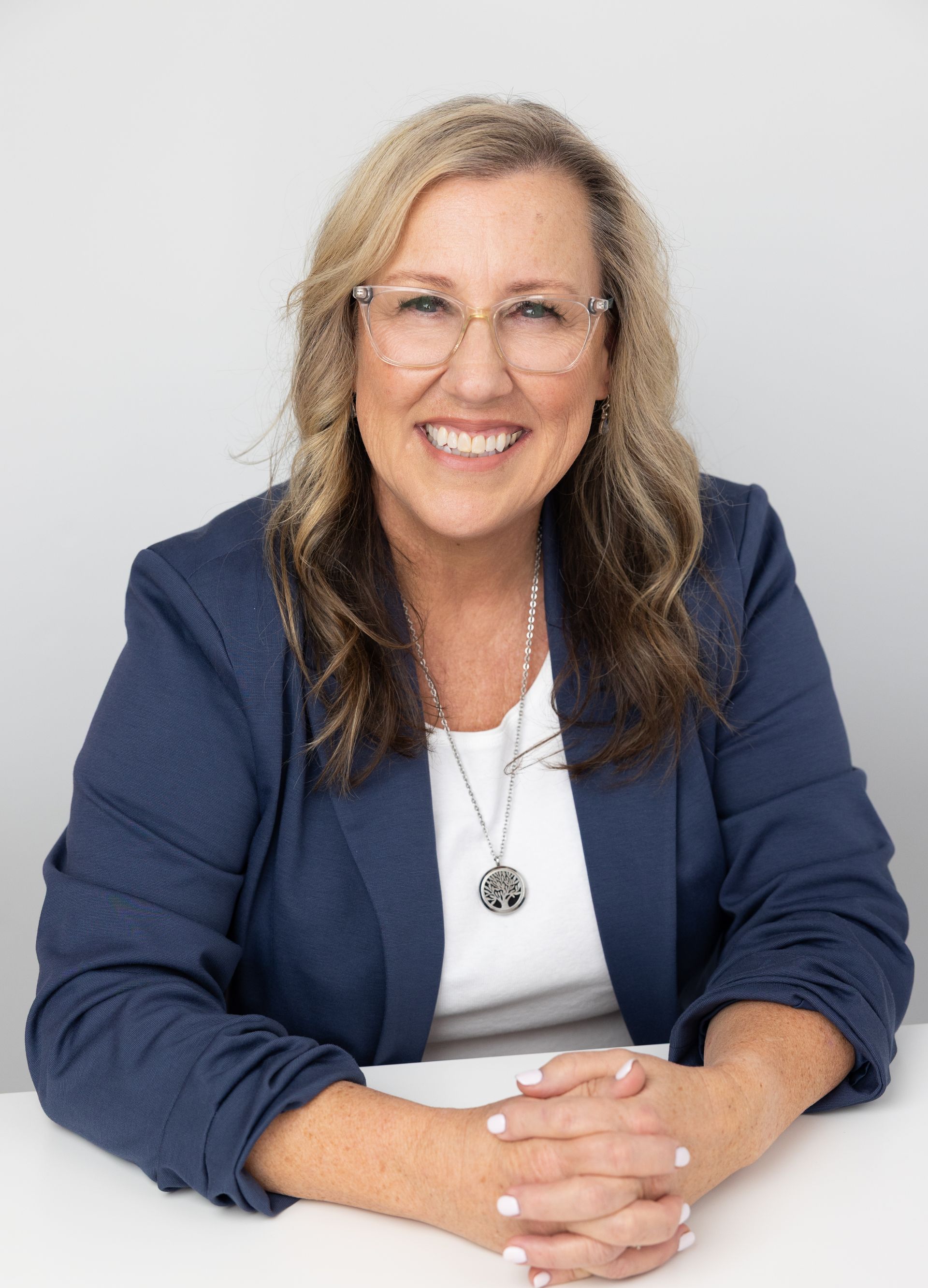 Smiling person in blue blazer seated at a table with hands clasped against a light background