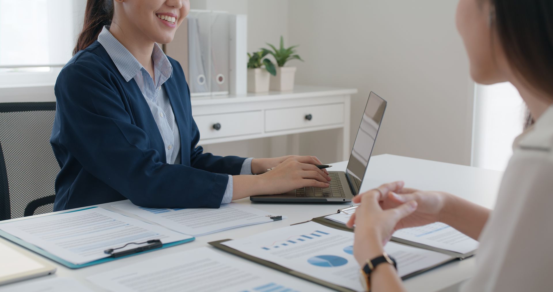 Two people discuss documents at a white office desk with a laptop and chart papers.