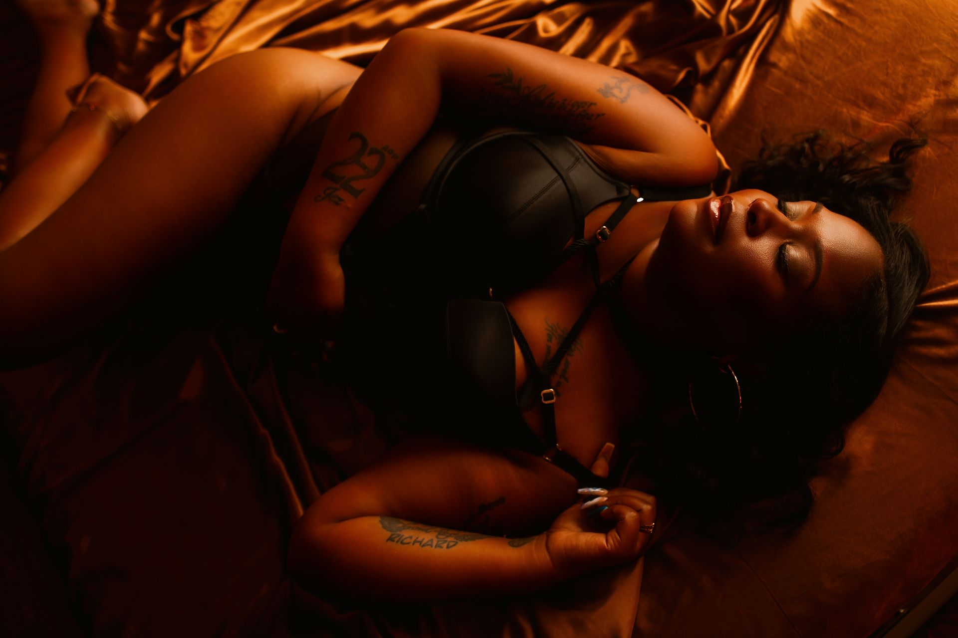 Person reclining on a bed in warm, dim orange light, wearing black lingerie and lying on a red blanket