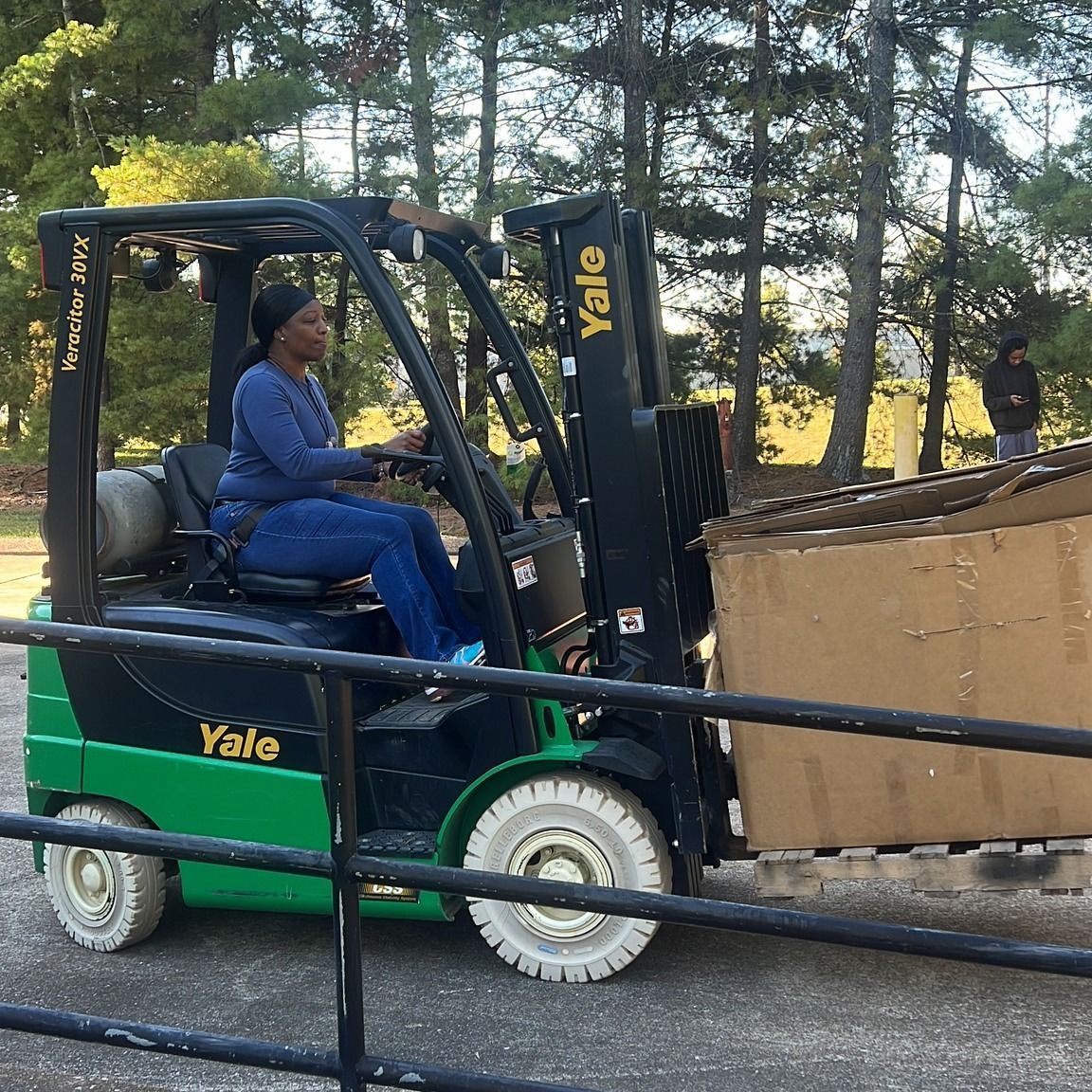 Woman operating a Yale forklift, carrying a pallet of boxes outdoors. Green, black, and white machinery.