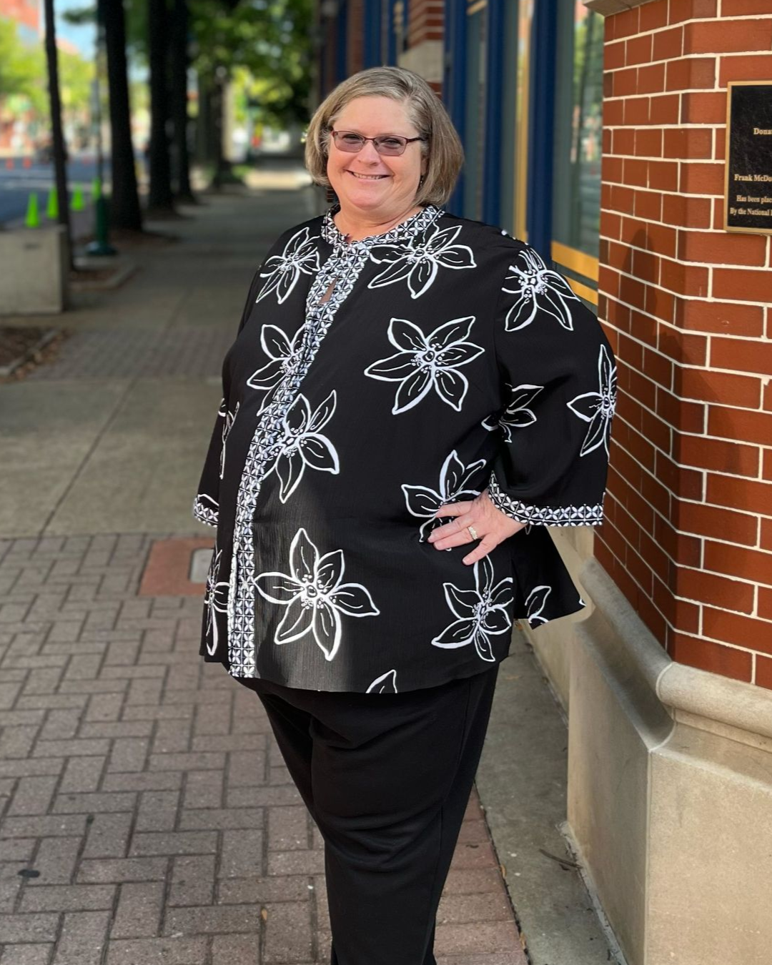Woman in black floral jacket and pants stands on a brick sidewalk next to a building.