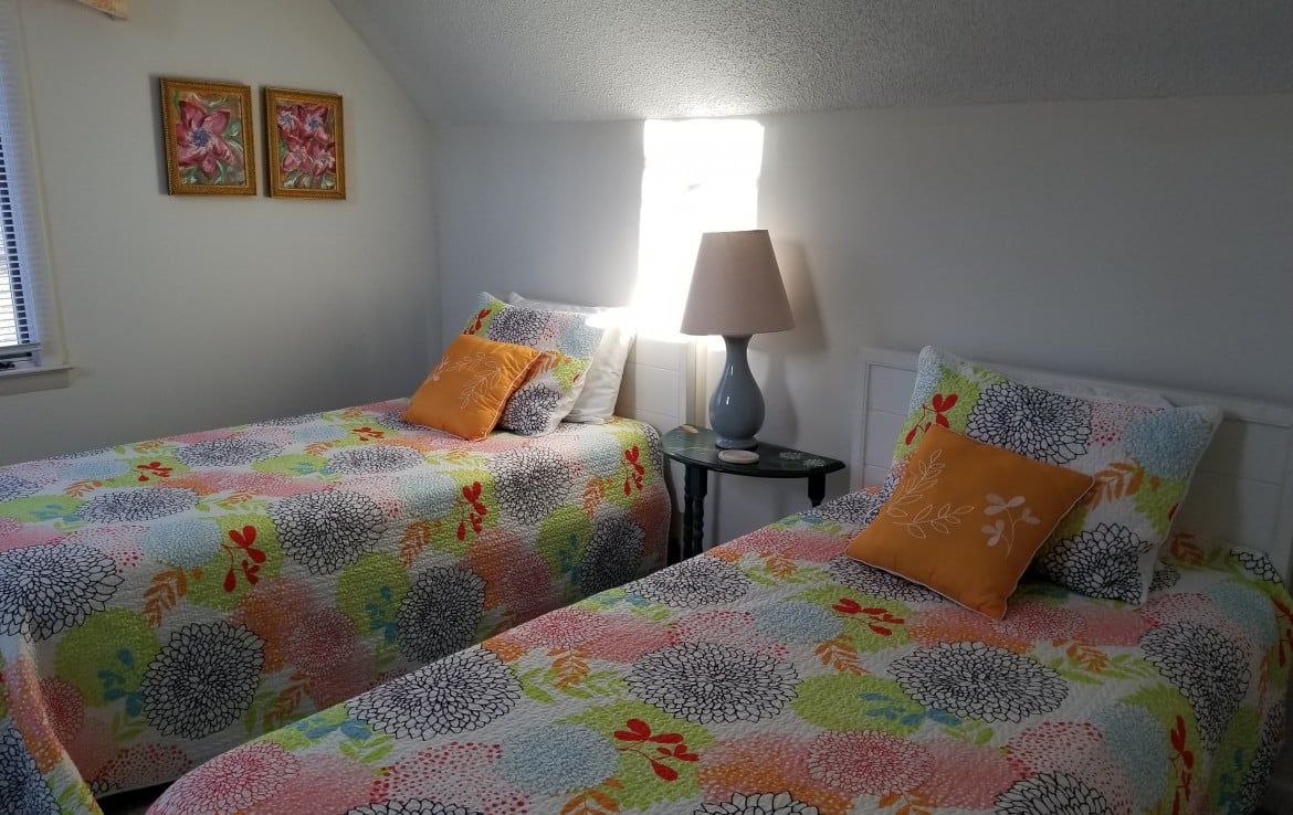 Two twin beds in a bright room with colorful patterned quilts, orange pillows, and a bedside lamp.