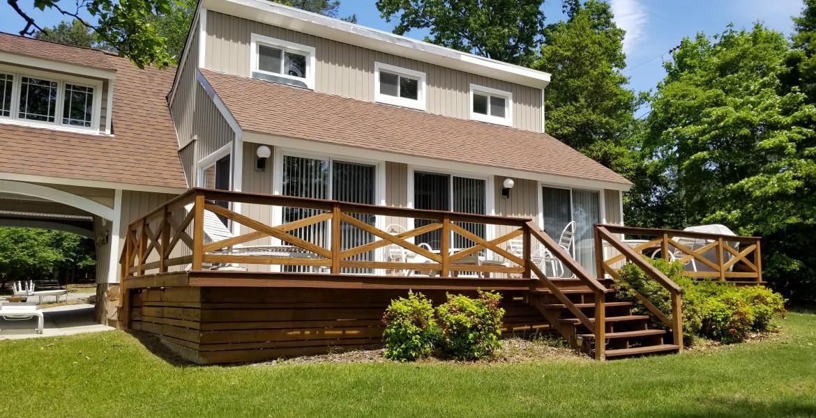 Beige house with a wooden deck and brown roof, surrounded by green grass and trees.