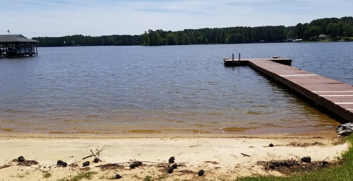 Sandy shoreline leading to a lake with a wooden dock. Trees line the far shore under a blue sky.