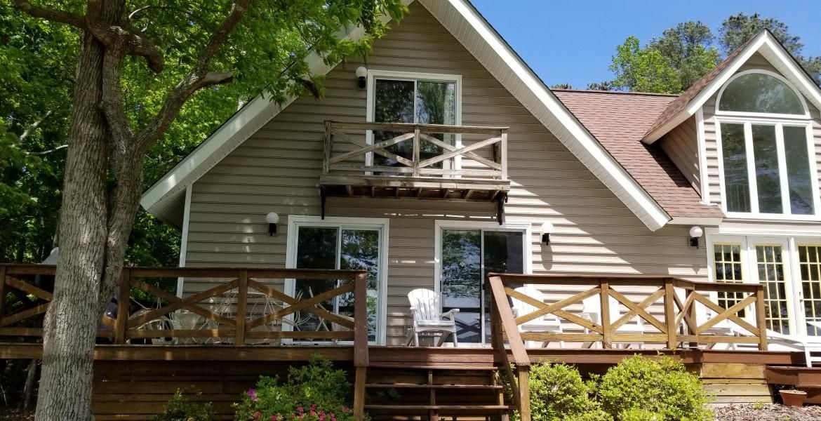 Two-story house with wooden deck and balcony. Beige siding, brown roof, and large windows. Sunny day.