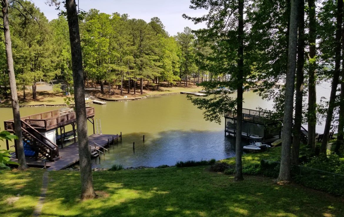 Lakeside view with docks, trees, and water. Green grass in foreground, trees frame the lake. Sunny day.