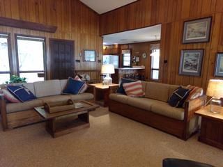 Cozy living room with wood paneling, two sofas with pillows, coffee table, and open doorway to kitchen.