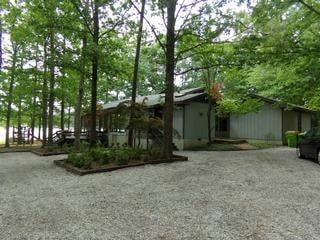 A one-story building with a gray exterior sits nestled among tall trees. A gravel driveway leads up to the house.