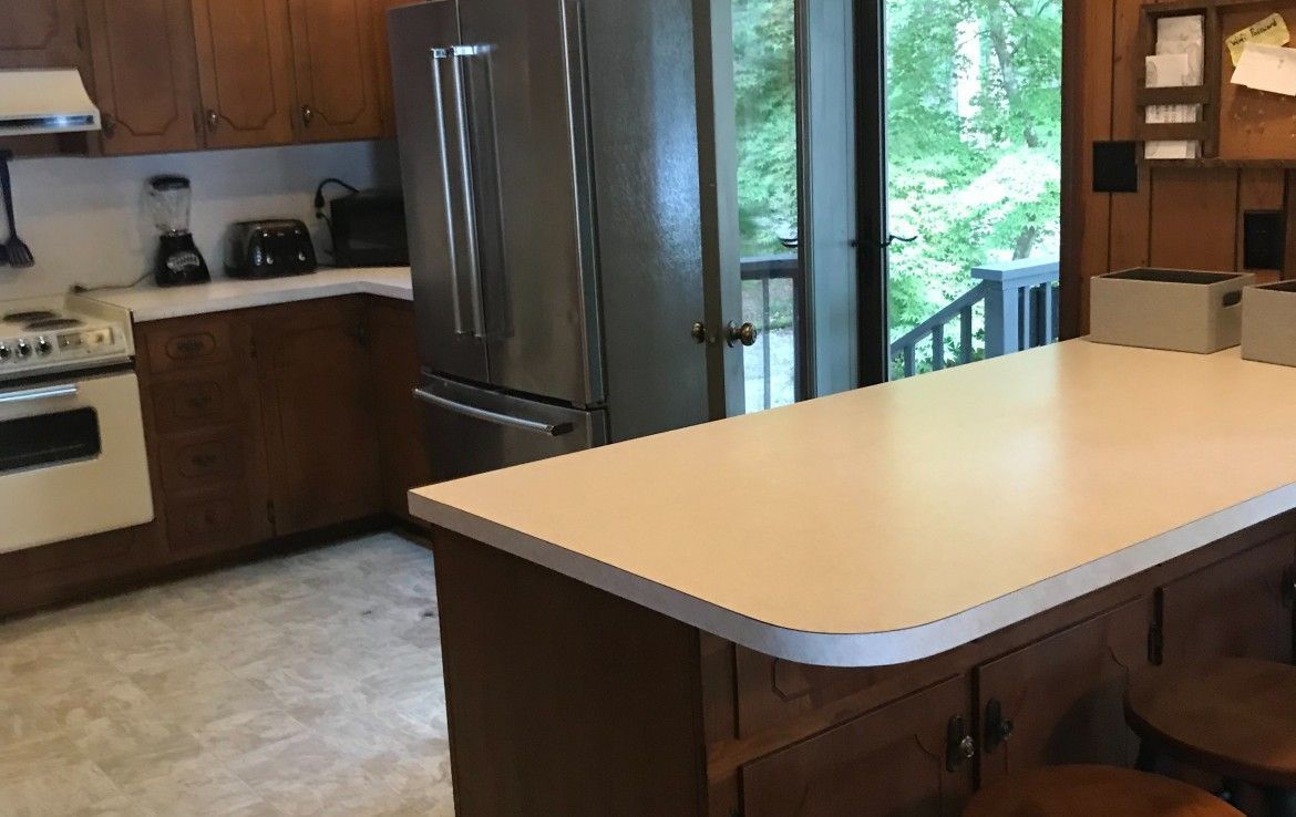 Kitchen with light-colored countertops, stainless steel fridge, and open door to a deck with trees.