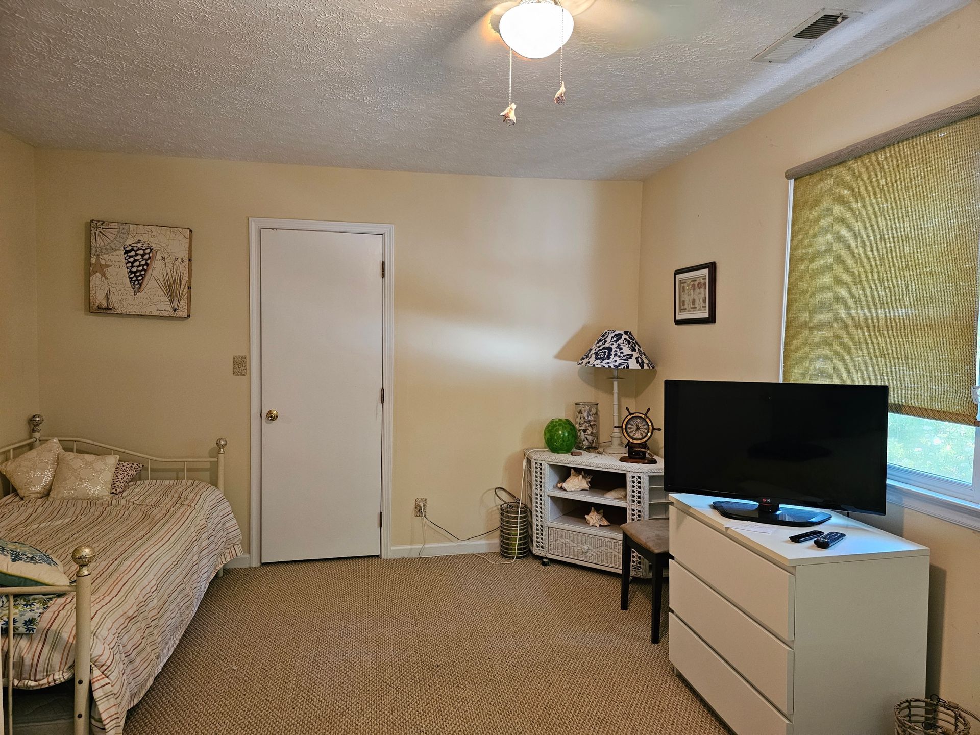 Bedroom with bed, door, chest of drawers with TV, window with blind, side table. Light yellow walls, patterned carpet.