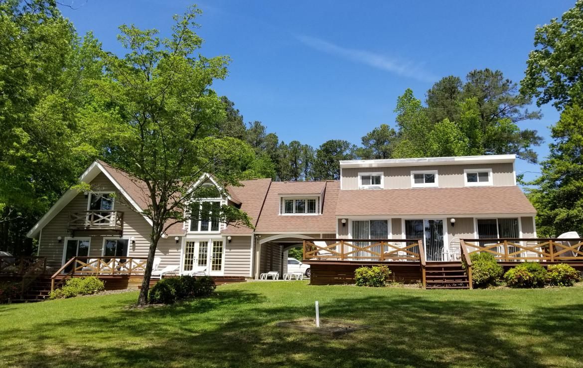 A two-story home with a wooden deck and green lawn against a background of trees and a blue sky.