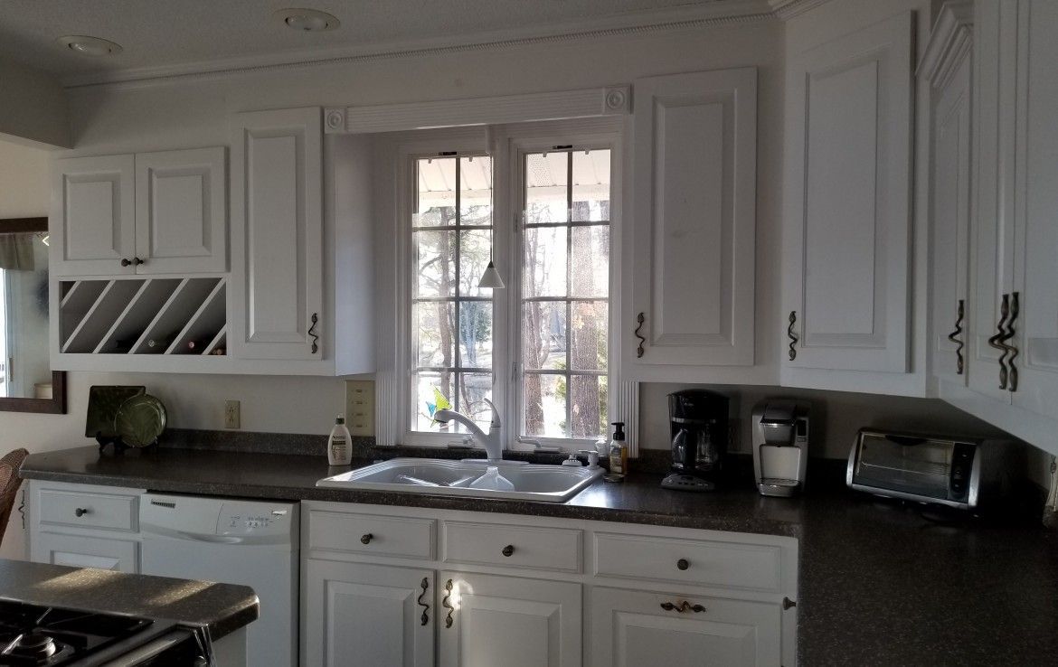 White kitchen with dark countertops, cabinets, wine rack, and sink under a window.