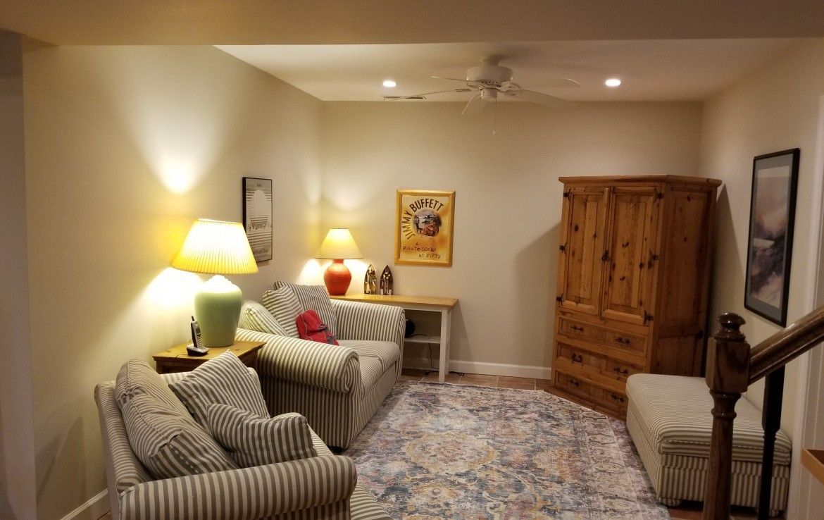 Cozy basement seating area with chairs, rug, cabinet, and side table; lit by lamps and ceiling lights.