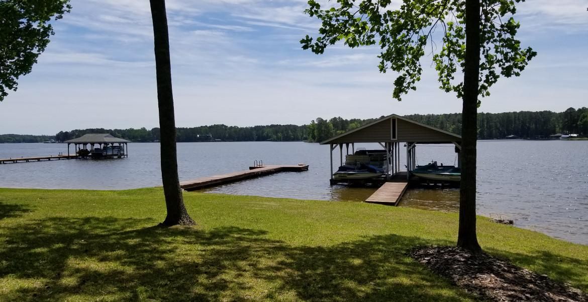 Lakeside scene with docks, boats under a covered slip, and a grassy shoreline under a blue sky.