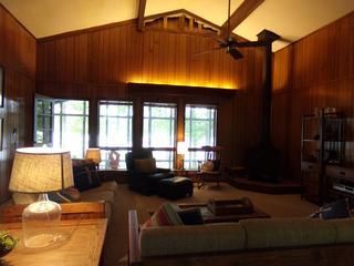 Cozy wood-paneled living room with large windows, fireplace, and ceiling fan; furnished with seating and lamps.
