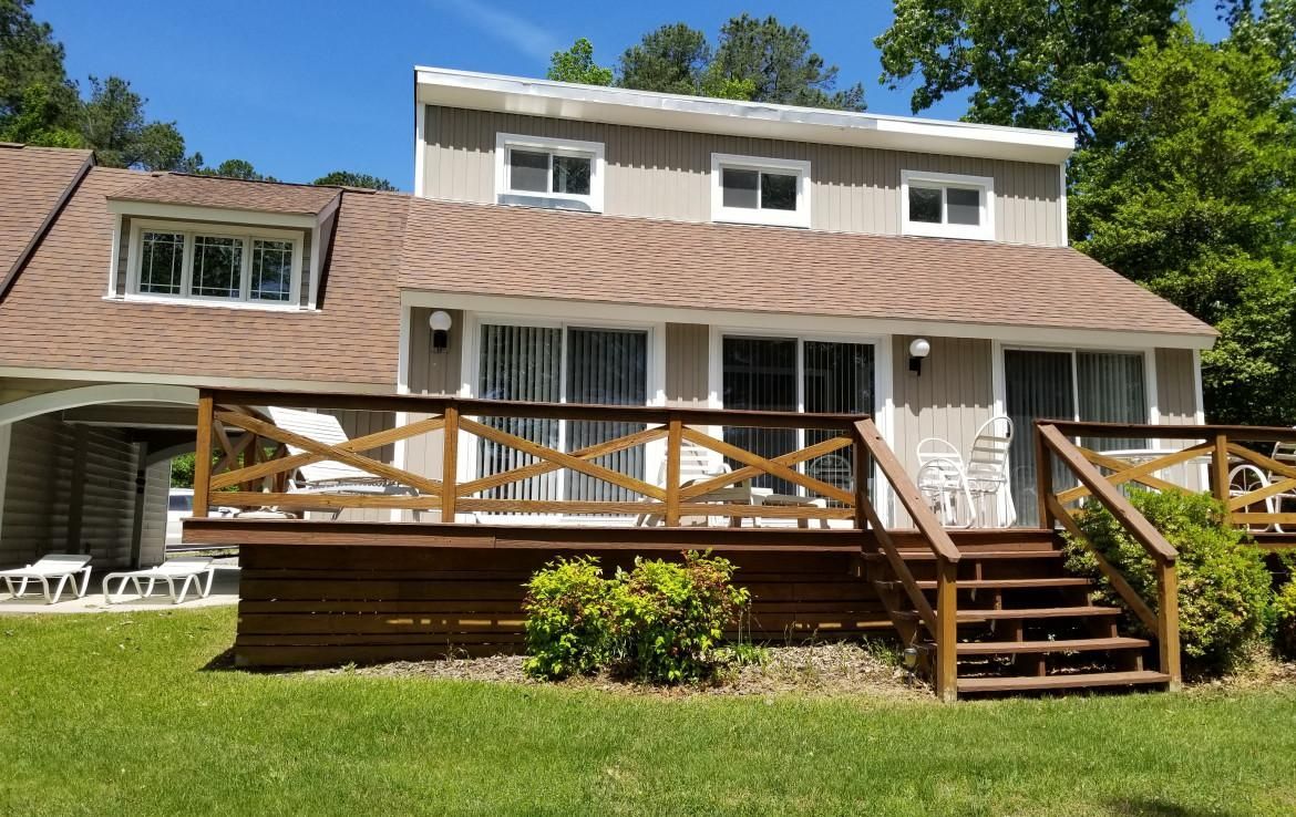 House with brown roof, deck, and steps; beige siding; green lawn; sunny day.