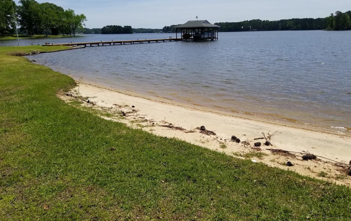 Lakeside view with grassy shore, sandy beach, and a covered dock. Blue water, blue sky.