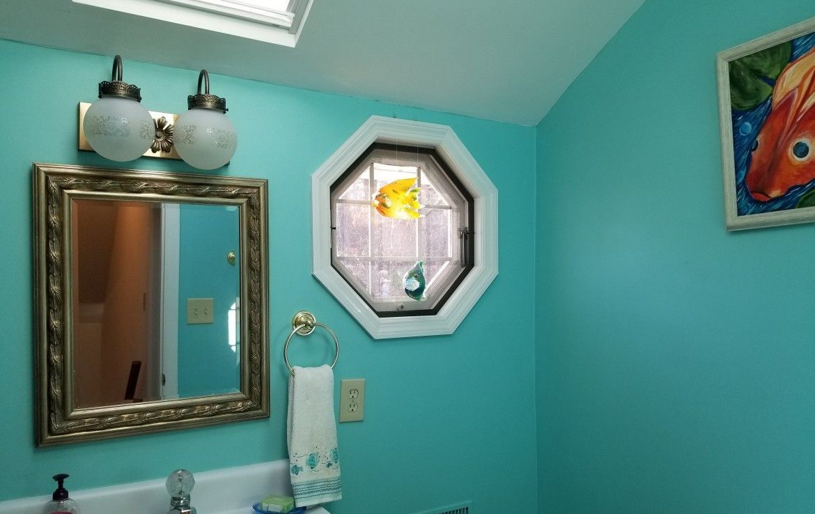 Teal bathroom with mirror, sconces, octagonal window, towel ring, and fish art.