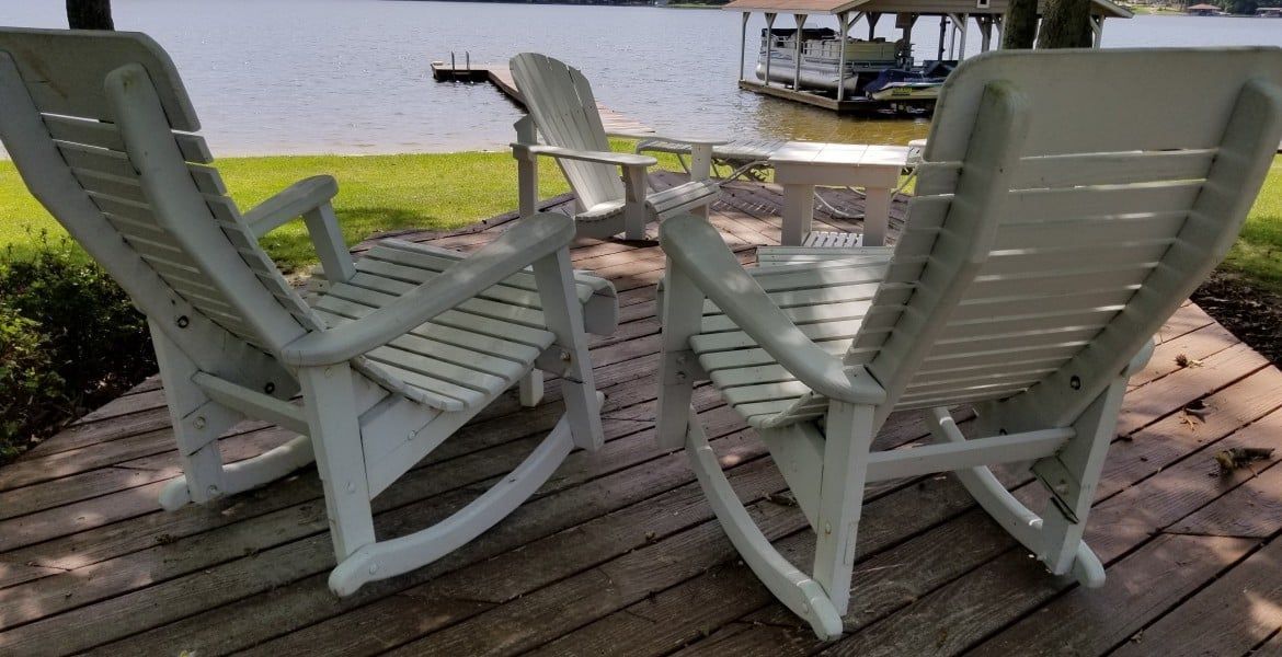 Two white rocking chairs on a deck, facing a lake with a dock and boathouse.