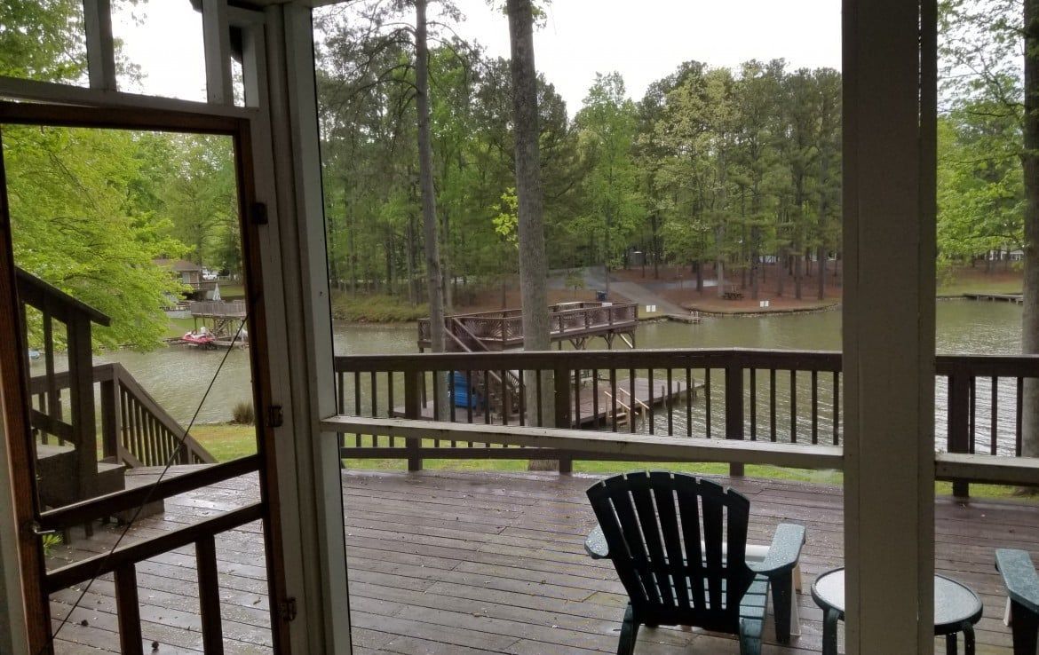 View from a covered porch to a lake with a wooden dock and trees, with a chair and table visible.