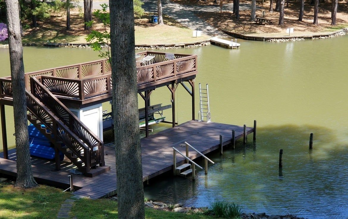 Wooden dock with stairs and a small upper deck on a lake. Trees surround the water's edge.