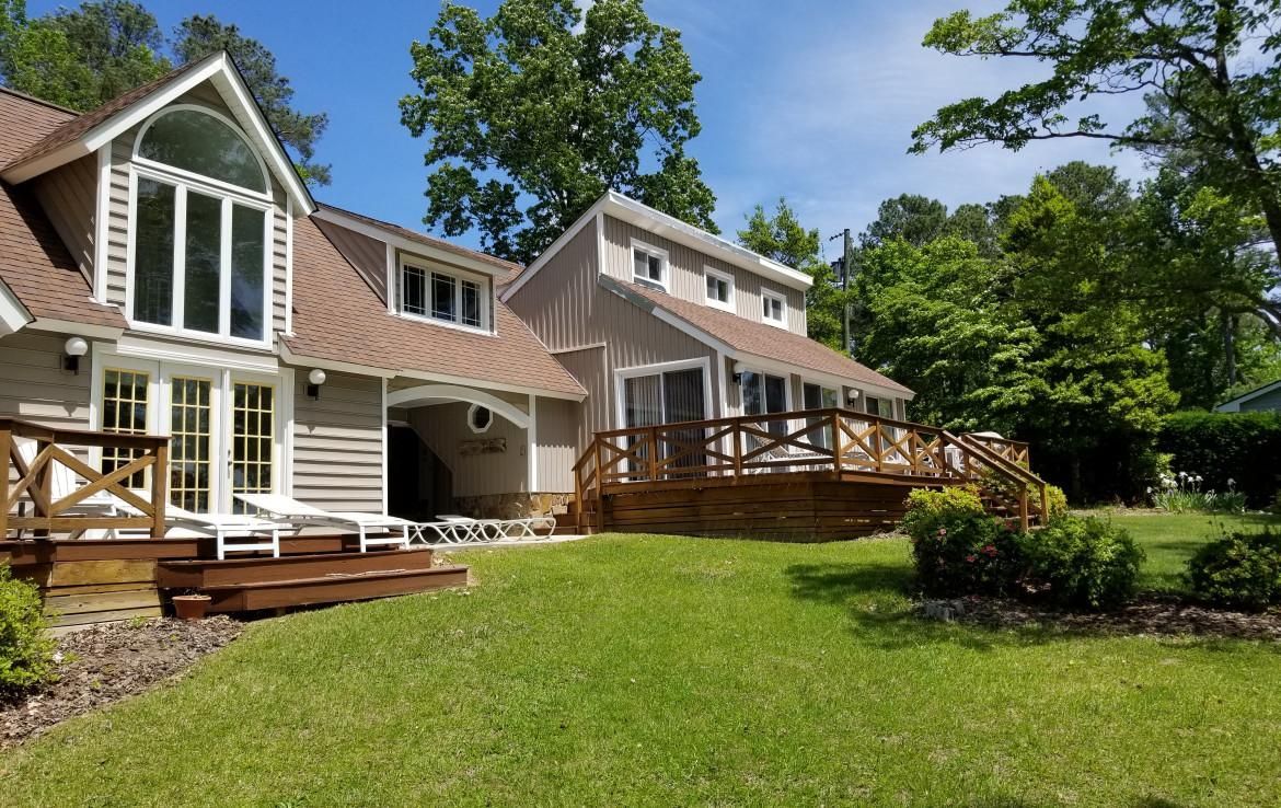 Beige house with a large window, wooden deck, and green lawn under a sunny sky.