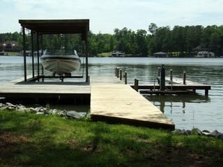 Boat on a lift under a canopy on a wooden dock extending into a lake. Green grass in foreground, trees in the background.
