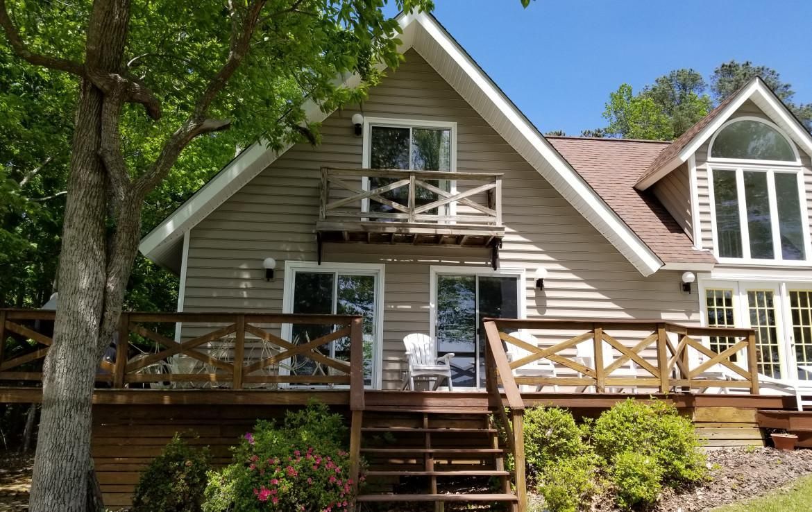 Tan A-frame house with brown deck and balcony, set against a blue sky, partially shaded by a tree.