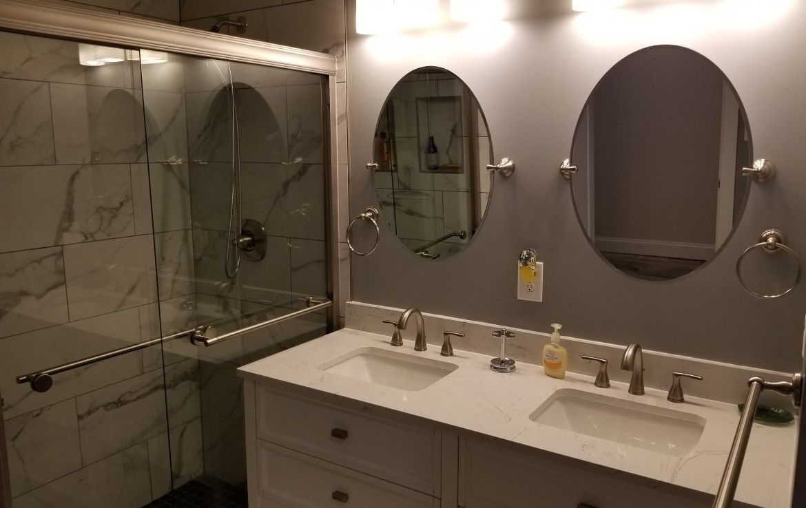 Bathroom with white vanity, marble-tiled shower, two oval mirrors, and brushed nickel fixtures.