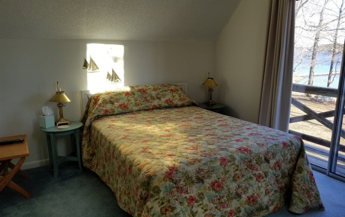 Bedroom with a floral patterned comforter on a bed, side tables, window, and view of trees and water.