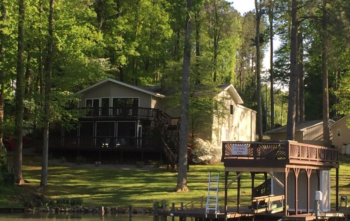 Cottage on a lake with a wooden dock and boathouse. Green trees surround the house.