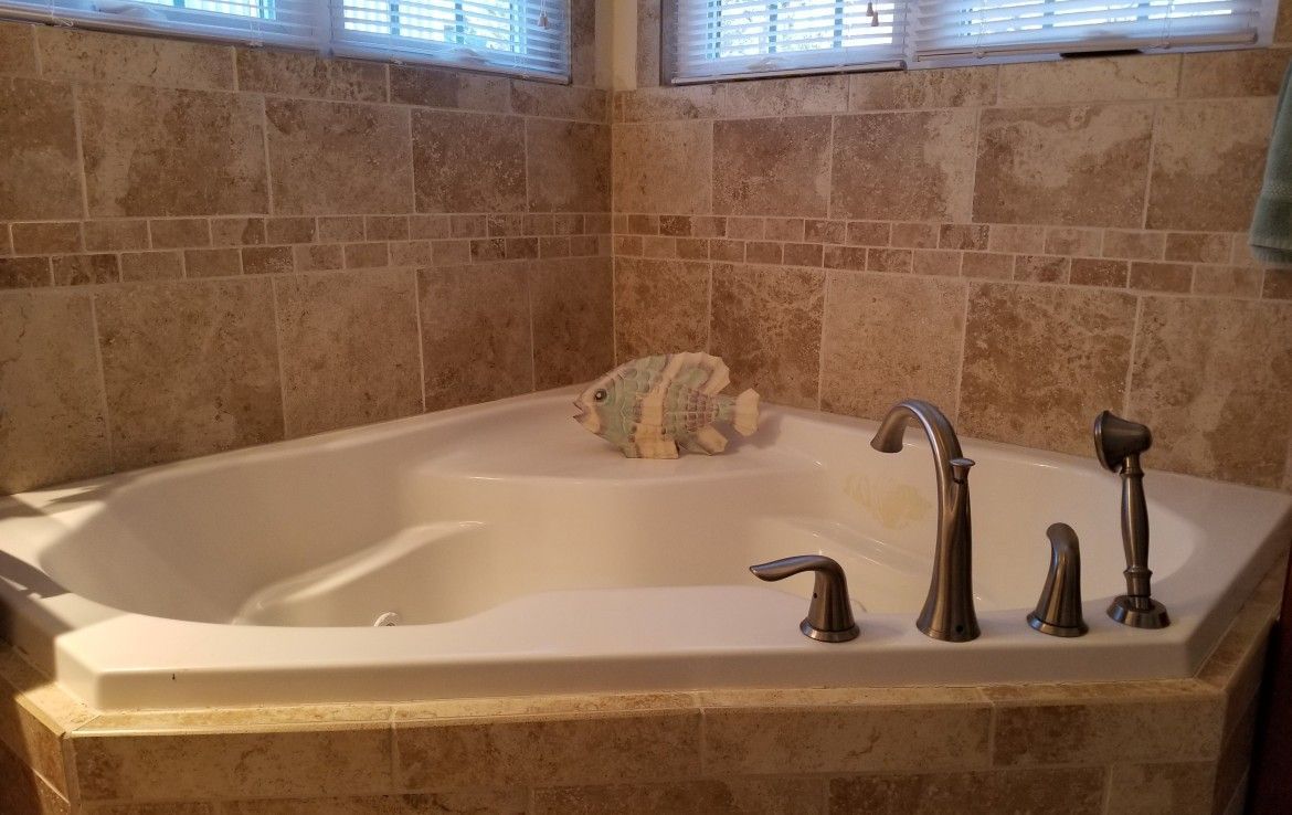 Corner bathtub surrounded by beige tile. Brushed nickel faucet. Towel on ledge.
