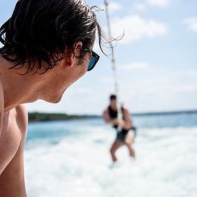 A shirtless man wearing sunglasses is watching a man wakeboard in the water.